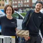 En kvinnlig och en manlig låssmed på en gata i Trollhättans centrum, bärandes på en stege och en låda från ASSA Abloy. I bakgrunden syns SafeTeams servicebilar.