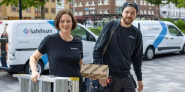 En kvinnlig och en manlig låssmed på en gata i Trollhättans centrum, bärandes på en stege och en låda från ASSA Abloy. I bakgrunden syns SafeTeams servicebilar.