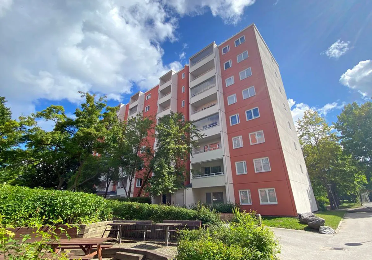Fastighet hos Botkyrkabyggen, ett flerfamiljshus med grönska och sittbänkar i förgrunden.