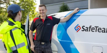 Säkerhetstekniker från SafeTeam samtalar med fastighetsskötare i gul varseljacka. De båda står vid SafeTeams servicebil, utanför en entré till ett flerfamiljshus.