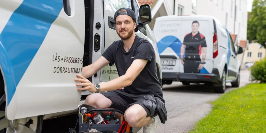 En porttelefonmontör från SafeTeam som sitter på huk bredvid sin servicebil, med en verktygslåda framför sig. I bakgrunden syns ett boendekvarter med lägenhetshus och ytterligare en SafeTeam-bil.