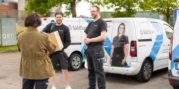 Två säkerhetstekniker från SafeTeam samtalar med en kvinna från en brf på en parkeringsplats. Bakom dem syns flera SafeTeam-bilar.