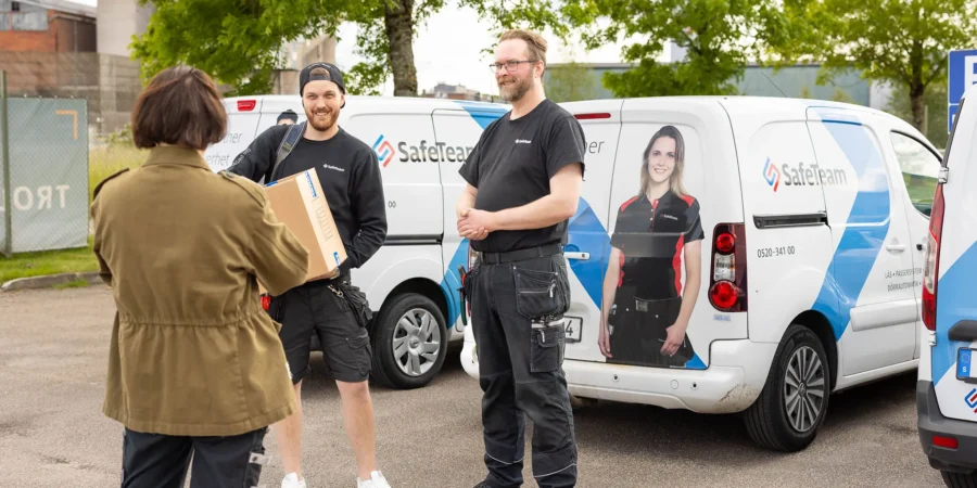 Två säkerhetstekniker från SafeTeam samtalar med en kvinna från en brf på en parkeringsplats. Bakom dem syns flera SafeTeam-bilar.
