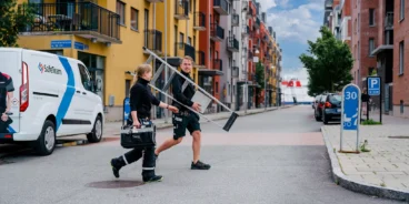 Två säkerhetstekniker passerar över en gata kantad av lägenhetshus i varma färger, med stege och verktygslåda i händerna.
