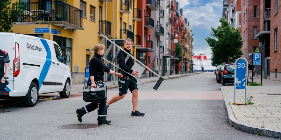 Två säkerhetstekniker passerar över en gata kantad av lägenhetshus i varma färger, med stege och verktygslåda i händerna.